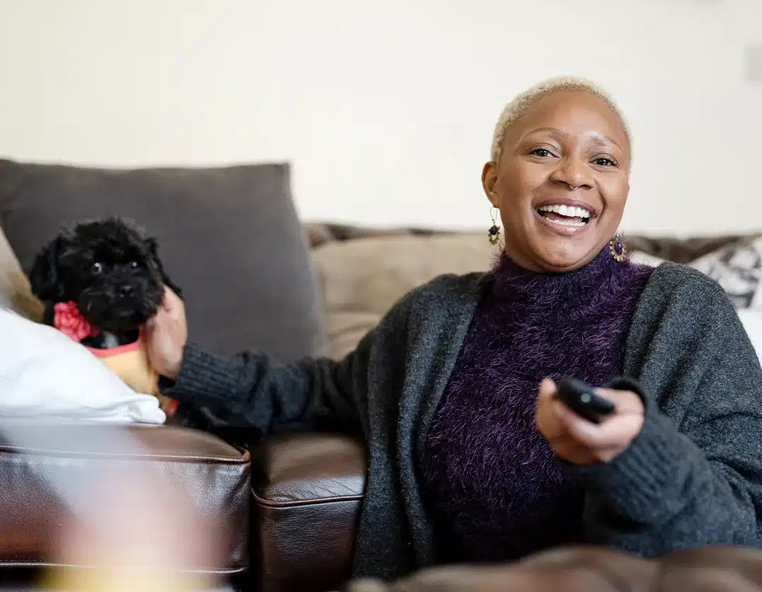 Black woman sitting on the couch, smiling, while holding out a TV remote